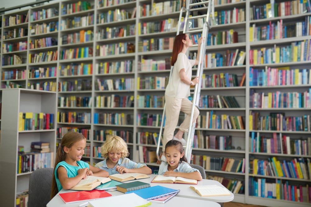 library-joyful-primary-school-children-reading-table-young-long-haired-woman-stairs-near-bookshelf-library